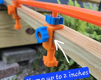 May include: Orange and blue toy track clamps, designed to secure a track to a wooden surface. The clamps can secure up to 2 inches. The track is orange, and the clamps have blue and orange components. The image shows a close-up of the clamp and track.