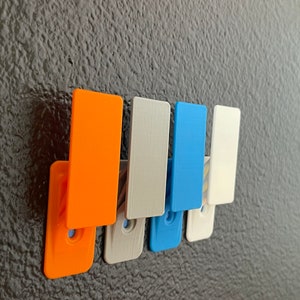 Pode incluir: Cinco ganchos de parede em diferentes cores: laranja, branco, azul, branco e branco. Os ganchos estão montados em uma parede cinza escura.