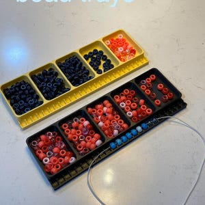 May include: Two bead trays, one yellow and one black, filled with colorful beads. The yellow tray holds black and orange beads. The black tray contains red, pink, and blue beads. The text "bead trays" is visible.