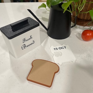 May include: White ceramic bread box with a black lid and the words "Fresh Bread" written in black script. A white plastic bread tag with the text "15 OCT" is on a white countertop. A brown plastic bread slice is next to the tag.