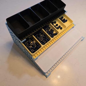 May include: A multi-compartment organizer with black and yellow trays, filled with small, dark blue objects. The organizer has a light blue frame and a white, textured surface. The top section has five rectangular compartments.