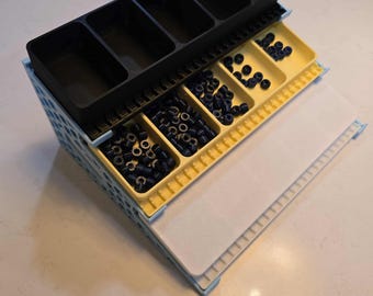 May include: A multi-compartment organizer with black and yellow trays, filled with small, dark blue objects. The organizer has a light blue frame and a white, textured surface. The top section has five rectangular compartments.
