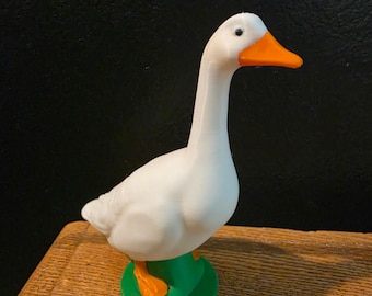 May include: A white goose figurine with an orange beak and feet, standing on a green base. The goose has a long, curved neck and is posed in a standing position. The figurine is likely made of plastic or a similar material.