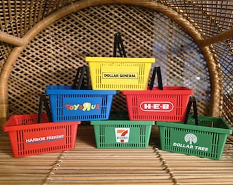 May include: A collection of miniature shopping baskets in various colors, including yellow, blue, red, and green. Each basket features a different store logo, such as Dollar General, Toys R Us, and H-E-B. The baskets have black handles.