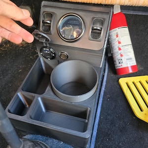 Può includere: Una console automobilistica grigio scuro con portabicchieri, vani portaoggetti e un manometro della pressione dell'airbag. Un estintore rosso e un attrezzo giallo sono visibili. La console ha anche una porta DC 5.0.