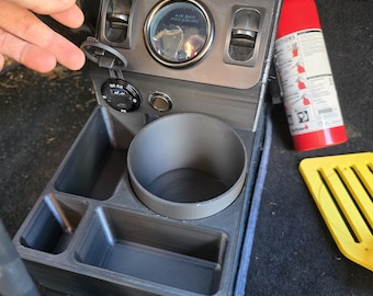 May include: A dark gray automotive console with a cup holder, storage compartments, and an air bag pressure gauge. A red fire extinguisher and a yellow tool are visible in the background. The console also has a DC 5.0 port.