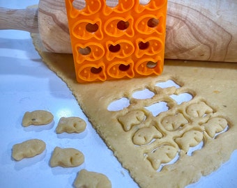 May include: Orange plastic cookie cutter shaped like a fish. The cutter is on top of a rolling pin and a sheet of dough. There are several fish-shaped cookies cut out of the dough.