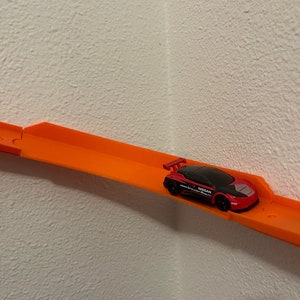 Può includere: Un'auto giocattolo rossa e nera con un logo "NISSAN" bianco e il testo "NISMO" sul lato, che guida su una pista di plastica arancione.
