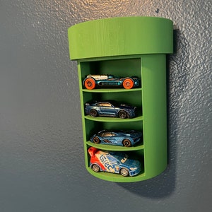 Op de afbeelding: Groene, pijp-vormige wandplank met vier planken om speelgoedauto's te tonen. De plank is gemaakt van hout en heeft een afgeronde bovenkant. Er staan vier speelgoedauto's op de planken, waaronder een blauwe auto met een rode en witte streep, een blauwe auto met een zwarte streep, een blauwe auto met een zilveren streep en een zilveren auto met rode wielen.