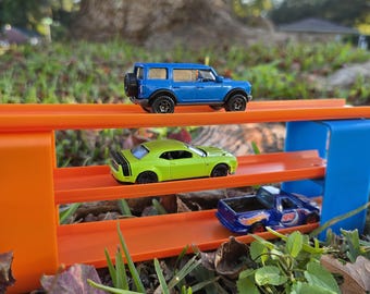 May include: A three-level orange plastic toy car track stand with blue side supports. A blue SUV toy car sits on the top level, a lime green sports car on the middle level, and a blue pickup truck toy car on the bottom level of the track.