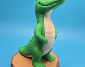 May include: A green crocodile figurine standing upright on a circular tan base. The crocodile has a smooth, light-colored belly, a textured back with small ridges, and a smiling expression with visible black eyes, presented in a standing pose.