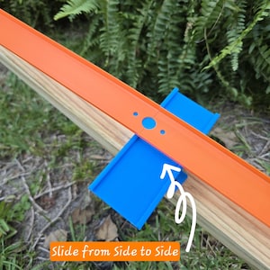 Könnte beinhalten: Eine orangefarbene Spielzeugrennstrecke mit blauen Stützen befindet sich auf einer Holzoberfläche. Der Text "Slide from Side to Side" ist auf dem Bild geschrieben. Die Strecke befindet sich vor einem Hintergrund aus grünem Laub.