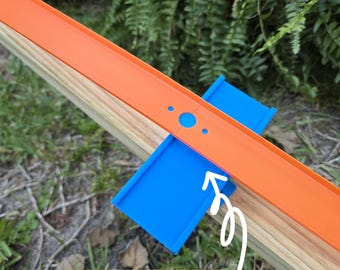 May include: An orange toy race track with blue supports is placed on a wooden surface. The text "Slide from Side to Side" is written on the image. The track is set against a backdrop of green foliage.