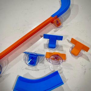 Könnte beinhalten: Ein Satz orangefarbener und blauer Kunststoff-Streckenstücke für ein Spielzeug-Autorennset. Die Streckenstücke umfassen eine gerade Strecke, eine Kurve und eine Rampe. Die Streckenstücke sind so konzipiert, dass sie mit Saugnäpfen an einer glatten Oberfläche befestigt werden können.