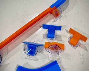 May include: A set of orange and blue plastic track pieces for a toy car racing set. The track pieces include straight track, curved track, and a ramp. The track pieces are designed to be attached to a smooth surface using suction cups.