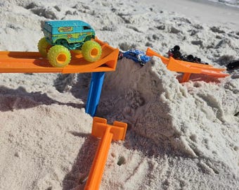 May include: A blue and green toy monster truck with the words "Monster Jam" on the side is driving on an orange plastic ramp. The ramp is set up on a white sandy beach with blue water in the background.
