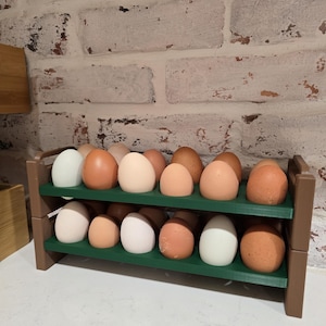 Puede incluir: Un estante para huevos de dos niveles hecho de madera marrón y verde, lleno de una variedad de huevos. Los huevos varían en color de blanco a marrón claro. El estante está sobre una encimera blanca contra una pared de ladrillo.
