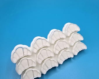 May include: A white plastic croissant-shaped cookie cutter with multiple cutouts. The cutter is on a blue background. The cutter is designed to cut out multiple croissants at once.