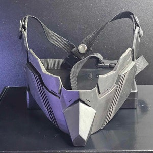 May include: A black plastic face mask with a geometric design and adjustable straps. The mask has a sharp, angular shape and a textured surface.