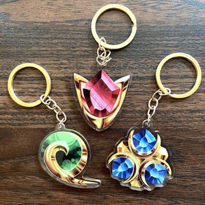 Peut inclure: Trois porte-clés en acrylique avec des anneaux et des chaînes en métal doré. L'un présente une forme de gemme rouge à facettes, un autre un motif circulaire vert tourbillonnant, et le troisième un groupe de trois gemmes bleues à facettes.