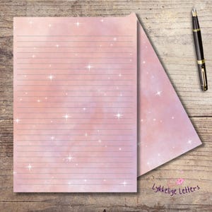 May include: A sheet of lined stationery paper with a soft pink and purple watercolor nebula background featuring scattered white stars, accompanied by a black fountain pen. The text Lykkelige Letters is printed in the bottom right corner.
