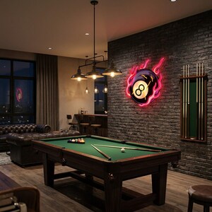 May include: A pool table with green felt, two cues, and a rack of balls sits in a room with a dark brick wall. A neon sign shaped like a flaming eight ball is mounted on the wall, and a wooden wall-mounted cue rack holds several pool cues.