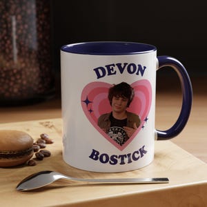 May include: A white ceramic mug with a dark blue interior and handle. The front features a pink heart graphic containing a photograph of Devon Bostick, surrounded by small star icons. The text DEVON BOSTICK is printed in dark blue above and below.
