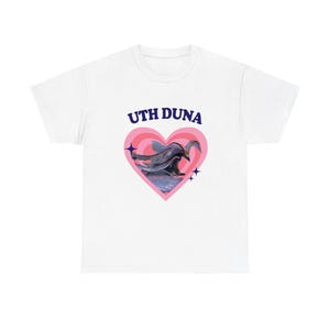 May include: A white short-sleeve t-shirt featuring a graphic print on the chest. The design includes the text UTH DUNA in dark blue letters above a pink heart shape containing an illustration of a bird in flight over water with small stars.
