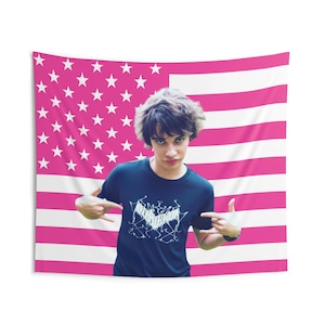 May include: A rectangular wall tapestry featuring a pink and white American flag pattern. Centered on the flag is a photograph of a person with dark hair wearing a dark blue t-shirt with a white graphic design on the front, making hand gestures.