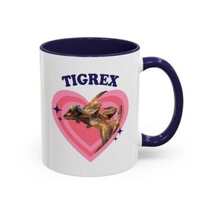 May include: A white ceramic mug with a dark blue interior and handle. The front features a pink heart graphic containing a brown and yellow Tigrex creature illustration, with the text TIGREX in dark blue letters above the heart and small dark blue stars.