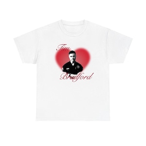May include: A white short-sleeve t-shirt featuring a central graphic of a man in a police uniform inside a red heart shape. The text Tim Bradford is printed in a red cursive script above and below the portrait of the man in uniform.