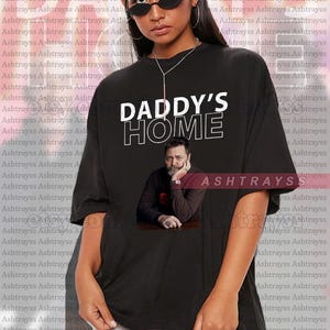 May include: Black unisex t-shirt featuring the text "DADDY'S HOME" in white. Underneath is a photo of a man. The shirt also has the word "ASHTRAYSS" printed on it.