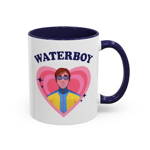 May include: White ceramic mug with a navy blue interior and handle. The mug features a graphic of a person inside a pink heart with the word "WATERBOY" above. The person is wearing blue goggles and a yellow and blue jacket.
