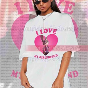 May include: White unisex t-shirt featuring a pink heart with Judy Hopps from Zootopia and the text "I LOVE MY GIRLFRIEND". The shirt is a casual style.