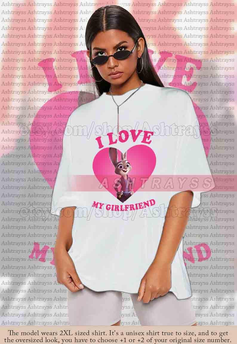 May include: White unisex t-shirt featuring a pink heart with Judy Hopps from Zootopia and the text "I LOVE MY GIRLFRIEND". The shirt is a casual style.