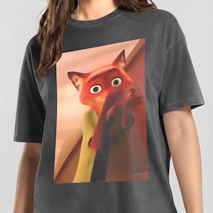 May include: Dark gray t-shirt featuring a cartoon image of a fox with orange fur and large eyes. The fox is the central focus, set against a blurred background. The shirt is a casual wear item.