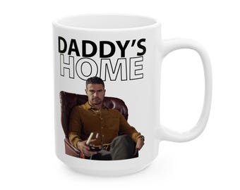 May include: A white ceramic mug featuring the black text DADDY'S HOME above a printed image of a person with dark hair wearing a mustard-colored button-down shirt, sitting in a dark brown leather armchair while holding a glass of red wine.
