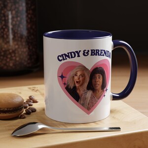 Peut inclure: Mug en céramique blanche avec intérieur et anse bleu marine. Le mug présente un motif de cœur rose avec deux visages de femmes et le texte "Cindy & Brenda" en bleu marine. Une cuillère et une pâtisserie sont au premier plan.