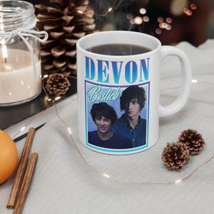 Puede incluir: Una taza de cerámica blanca con un estampado gráfico del actor Devon Bostick. El diseño incluye el nombre DEVON en letras azules en negrita en la parte superior, el nombre Bostick en una fuente cursiva azul claro y dos retratos del actor sobre un fondo azul.