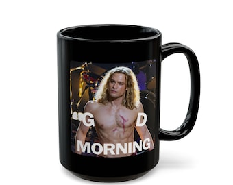 Puede incluir: Una taza de cerámica negra con la imagen impresa de una persona sin camisa, con cabello largo rubio y una cicatriz en el pecho. El texto "G D MORNING" está impreso en letras mayúsculas blancas sobre la imagen, que muestra instrumentos musicales.
