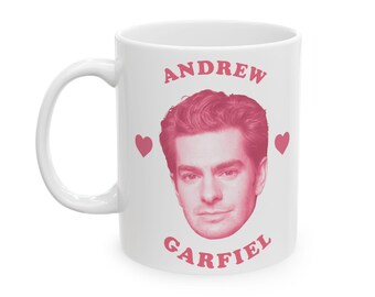 May include: White ceramic mug featuring a pink image of Andrew Garfield's face, with the name "ANDREW GARFIELD" in pink lettering. Two pink hearts flank the image. The mug has a white handle.