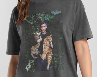 May include: A charcoal gray short-sleeve t-shirt featuring a graphic print of a person holding two cats, one tabby and one orange, against a dark green foliage background. The shirt has a crew neckline and a relaxed, boxy fit with short sleeves.