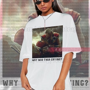 Puede incluir: Camiseta unisex blanca con un gr&aacute;fico de Thor sosteniendo a Deadpool, con el texto "WHY WAS THOR CRYING?". La marca "ASHTRAYSS" tambi&eacute;n est&aacute; impresa en la camiseta.