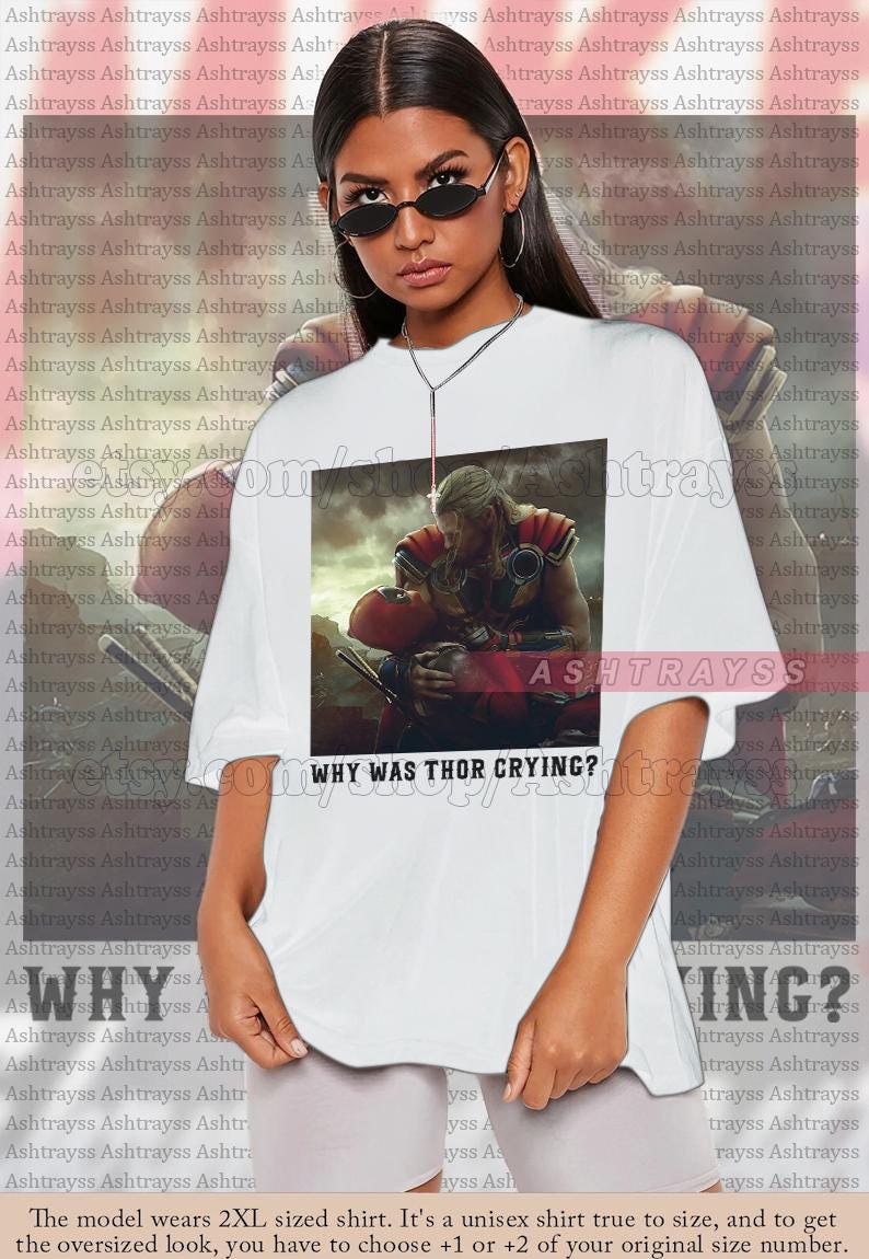 Puede incluir: Camiseta unisex blanca con un gr&aacute;fico de Thor sosteniendo a Deadpool, con el texto "WHY WAS THOR CRYING?". La marca "ASHTRAYSS" tambi&eacute;n est&aacute; impresa en la camiseta.
