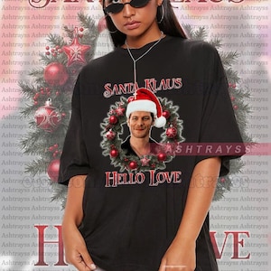 Op de afbeelding: Een zwart T-shirt met een kerstkransontwerp met een man die een kerstmanmuts draagt. De krans is versierd met rode en groene kerstballen. De tekst "Santa Klaus" en "Hello Love" staat op het shirt gedrukt.