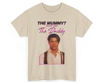 May include: Beige t-shirt with a graphic print of a man with the text "THE MUMMY? more like The Daddy" in pink.