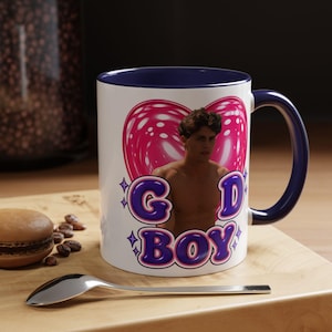 May include: A white ceramic mug with a dark blue interior and handle. The front features a graphic of a person in front of a pink heart, with the purple text "GOD BOY" below, accented by small purple star icons on a white background.