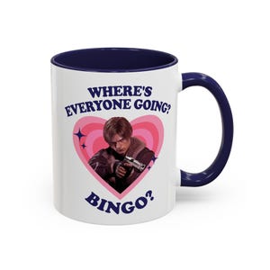 Può includere: Tazza in ceramica bianca con interno e manico blu navy. La tazza presenta una grafica di una persona che tiene una pistola all'interno di un cuore rosa con il testo "WHERE'S EVERYONE GOING? BINGO?"
