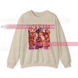Puede incluir: Sudadera beige con un gr&aacute;fico de varios zorros de dibujos animados y el texto "NICK WILD" en rojo. Los zorros est&aacute;n en varias poses y visten camisas moradas. La sudadera tiene mangas largas y cuello redondo.
