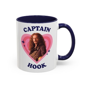 May include: A white ceramic mug with a dark blue interior and handle. The front features a pink heart graphic containing a portrait of Captain Hook, surrounded by small dark blue star icons. The text CAPTAIN HOOK is printed in dark blue.